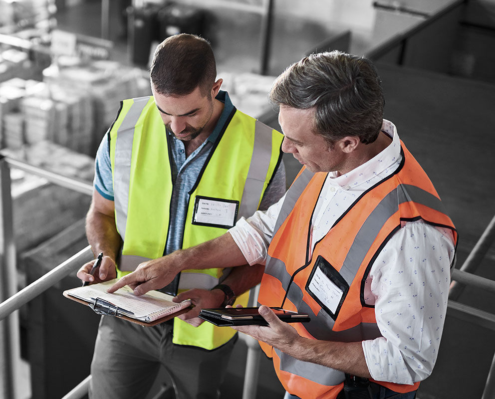 Professional employees wearing safety vests, looking at a clipboard together in a warehouse