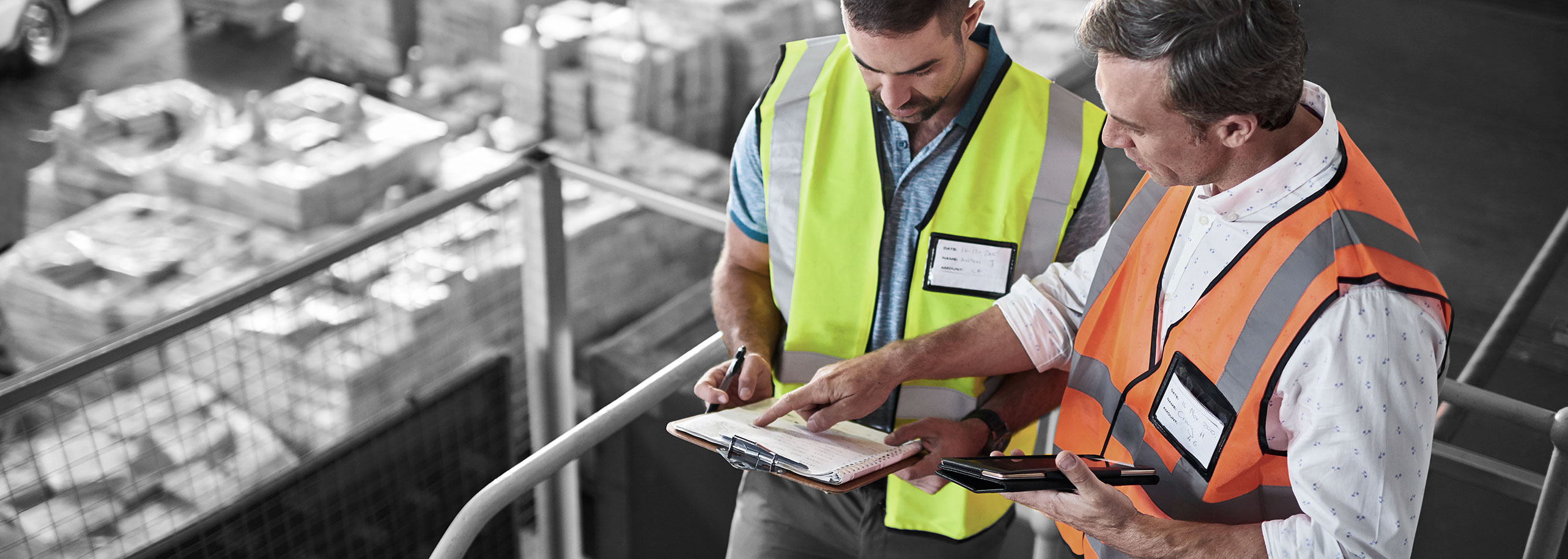 Professional employees wearing safety vests, looking at a clipboard together in a warehouse